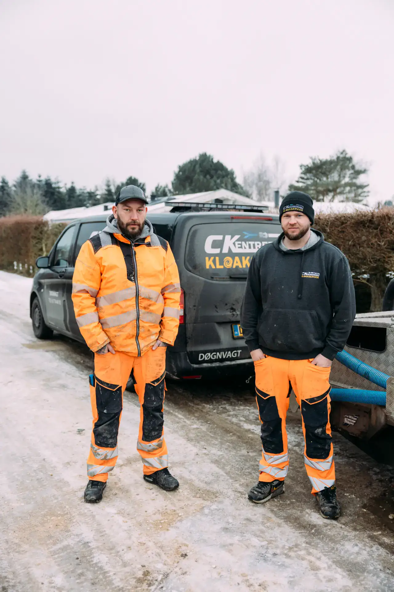 To kloakarbejdere står foran firmaets servicebil i udendørs miljø.