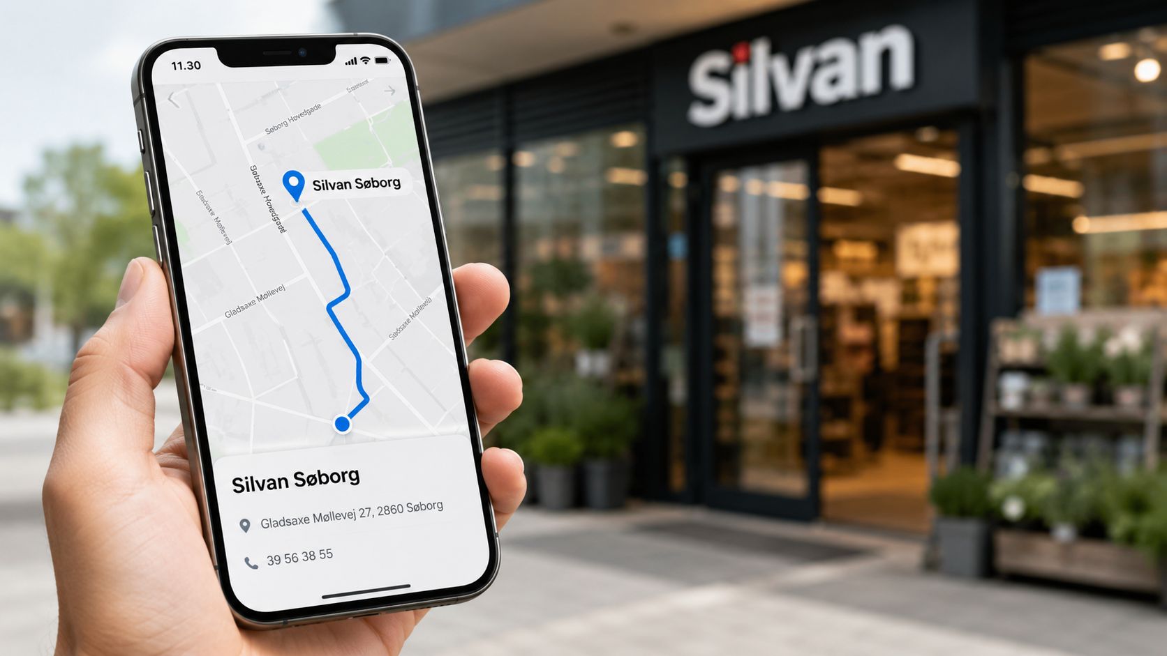En person holder en smartphone, der viser kort og kontaktoplysninger til Silvan Søborg-butikken på skærmen.